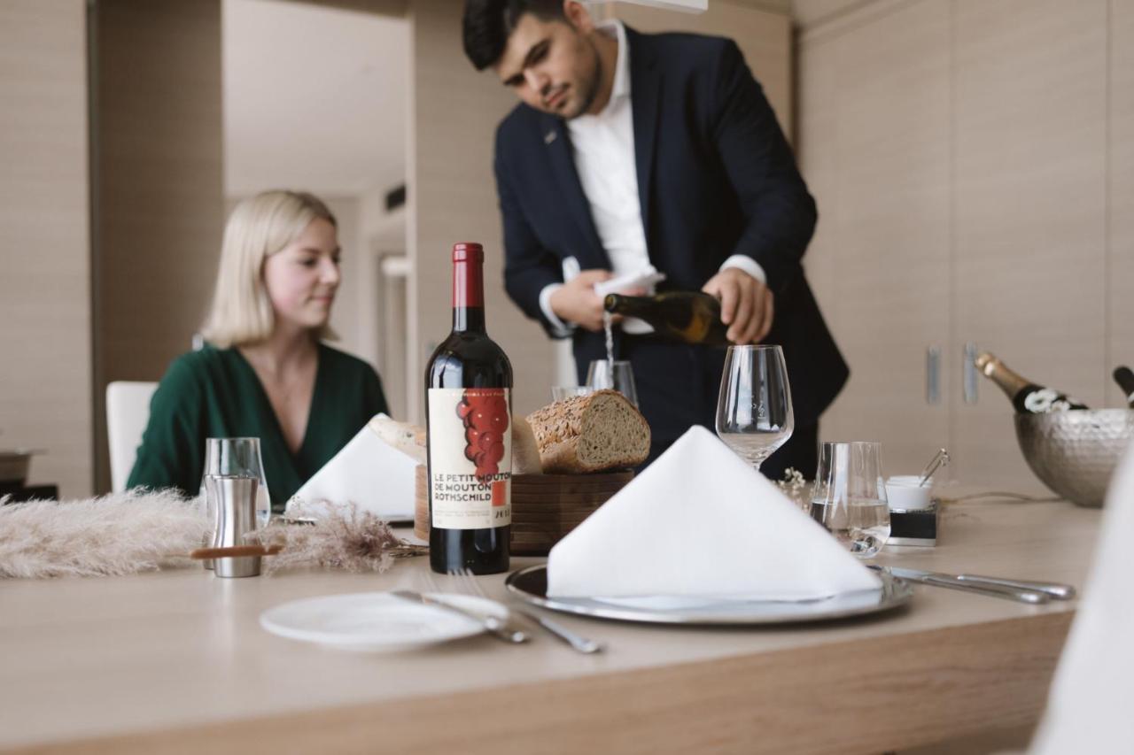 더 웨스틴 함부르크 함브루크 외부 사진 The photo shows a dining scene featuring a well-dressed man pouring wine into a glass while a woman sits at the table. The table is elegantly set with a folded napkin, wine glasses, and a bottle of red wine placed prominently. There is also a loaf of