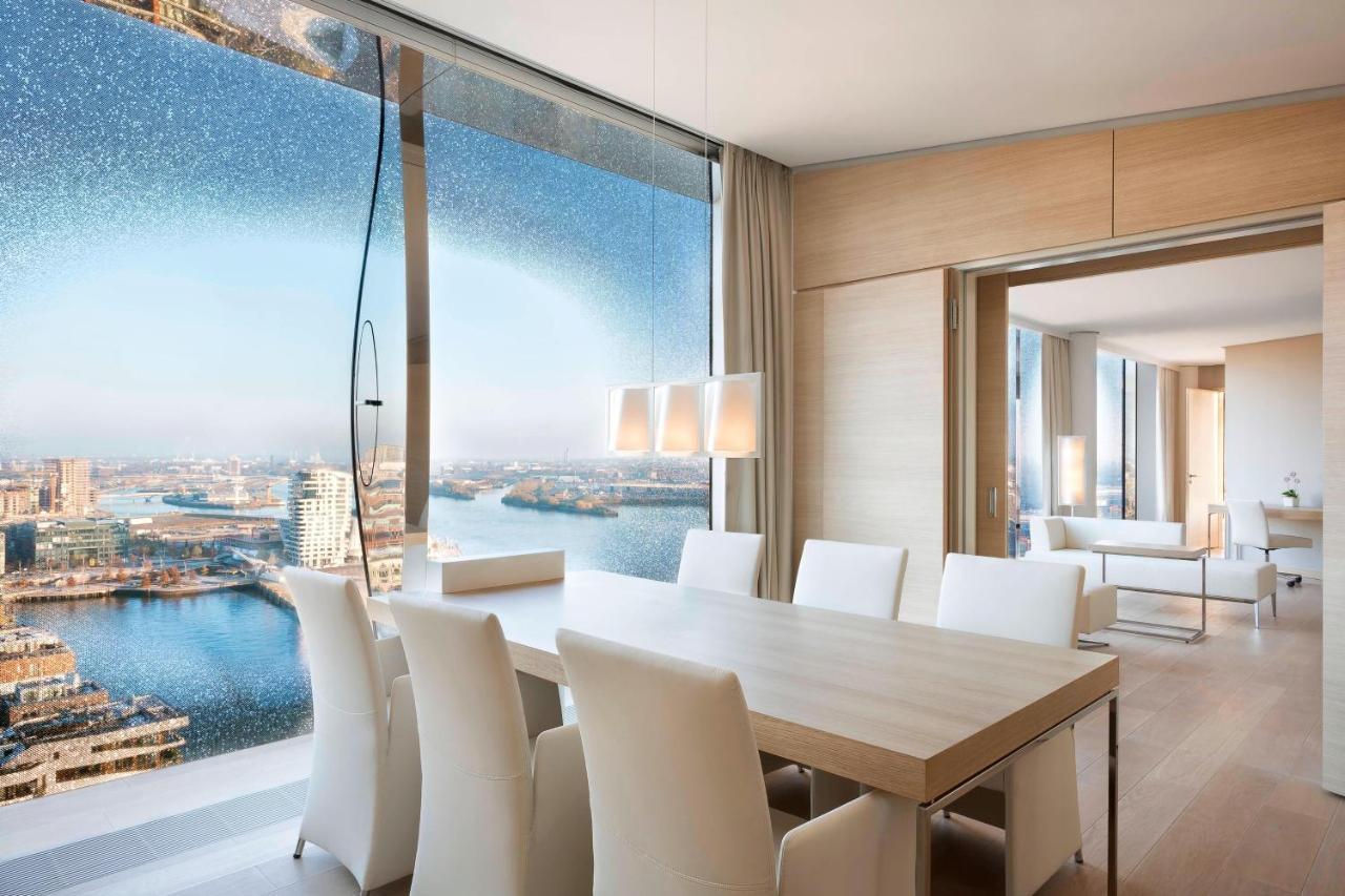 더 웨스틴 함부르크 함브루크 외부 사진 The photo shows a modern dining area with a large, light-colored table surrounded by white chairs. The space features a panoramic window that offers a stunning view of a body of water and a cityscape. The decor is minimalistic and elegant, with smoot
