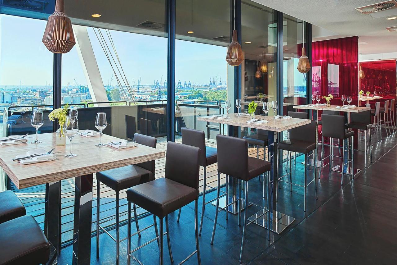 더 웨스틴 함부르크 함브루크 외부 사진 The photo depicts a modern restaurant interior featuring a spacious dining area with large windows that provide a view of the outside. There are several tall tables with a light-colored surface and metallic bases, accompanied by dark, sleek stools. T
