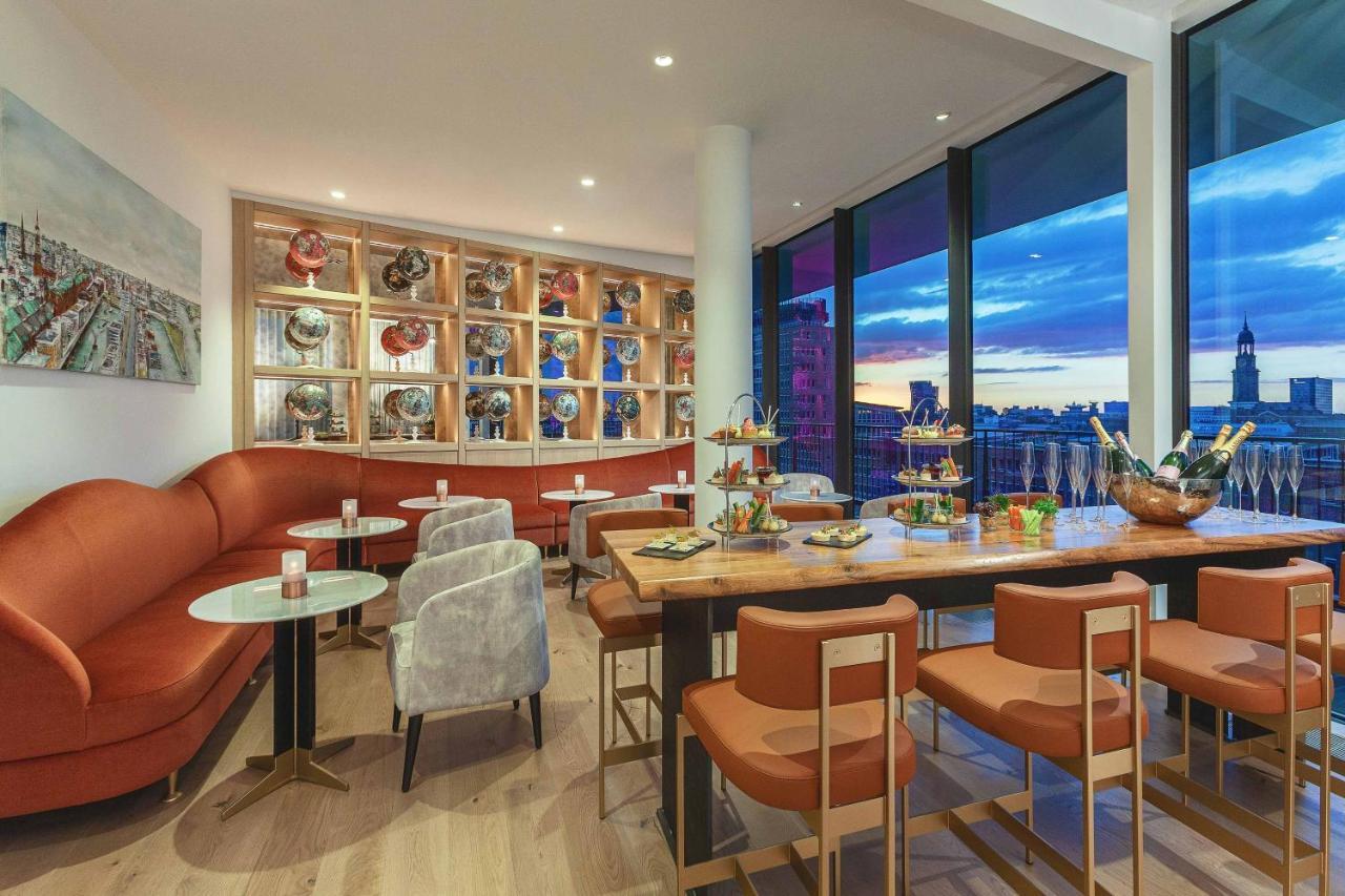 더 웨스틴 함부르크 함브루크 외부 사진 The photo shows a modern and stylish restaurant or café interior. It features a combination of comfortable seating, including a curved, orange sofa and gray chairs. The tables are round with light-colored tops, and there are several tiered stands dis