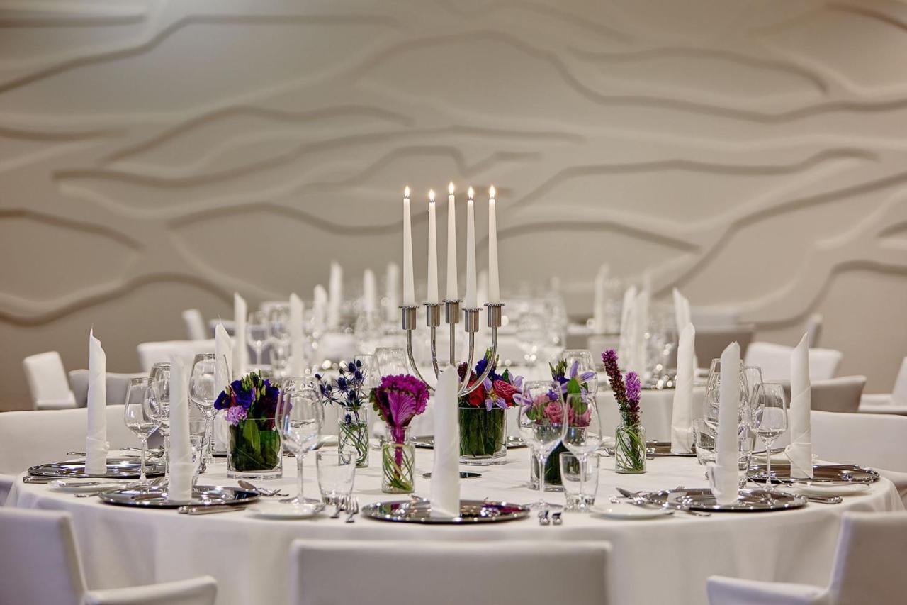 더 웨스틴 함부르크 함브루크 외부 사진 The photo depicts an elegantly set round dining table. In the center, there is a candelabra with several tall candles, providing a soft, warm light. Surrounding the candelabra are small glass vases filled with colorful flowers, adding a vibrant touch