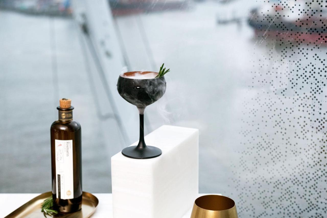 더 웨스틴 함부르크 함브루크 외부 사진 The photo features a stylish cocktail elegantly presented on a white pedestal. The cocktail is served in a sophisticated black stem glass, topped with a garnish of rosemary. Beside it, there is a dark glass bottle, likely containing a spirit or mixer