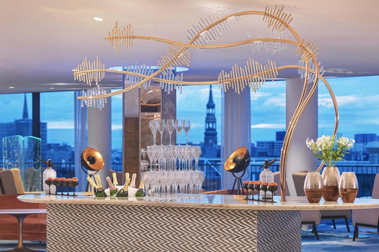 더 웨스틴 함부르크 함브루크 외부 사진 The photo depicts a stylish bar setup in a modern venue. The bar features an elegant design with a patterned front and a sleek countertop. Above the bar, there is a striking decorative element that resembles a stylized tree or wave, made from metalli