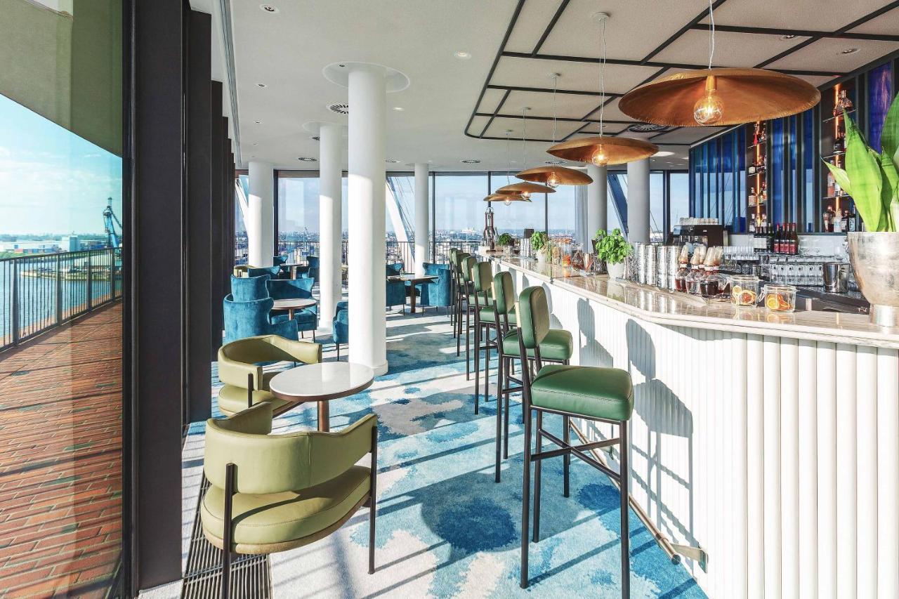 더 웨스틴 함부르크 함브루크 외부 사진 The photo depicts a modern bar or lounge area with a stylish design. There are high-backed green chairs surrounding small circular tables, arranged in an open space that has large windows showcasing a view of the outside. The bar area is sleek, featu
