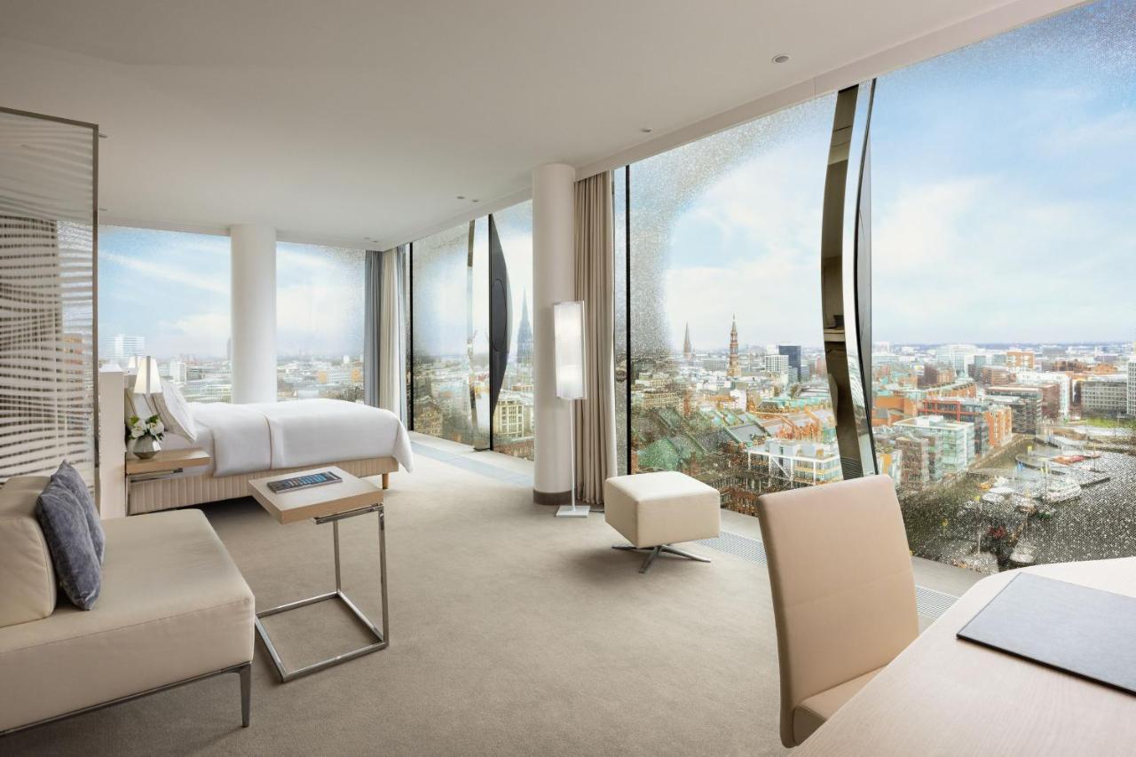 더 웨스틴 함부르크 함브루크 외부 사진 The photo shows a modern hotel room with large floor-to-ceiling windows that provide a panoramic view of a city landscape. The room features a minimalist design with neutral tones. There's a king-sized bed positioned in the center, a sleek desk and c