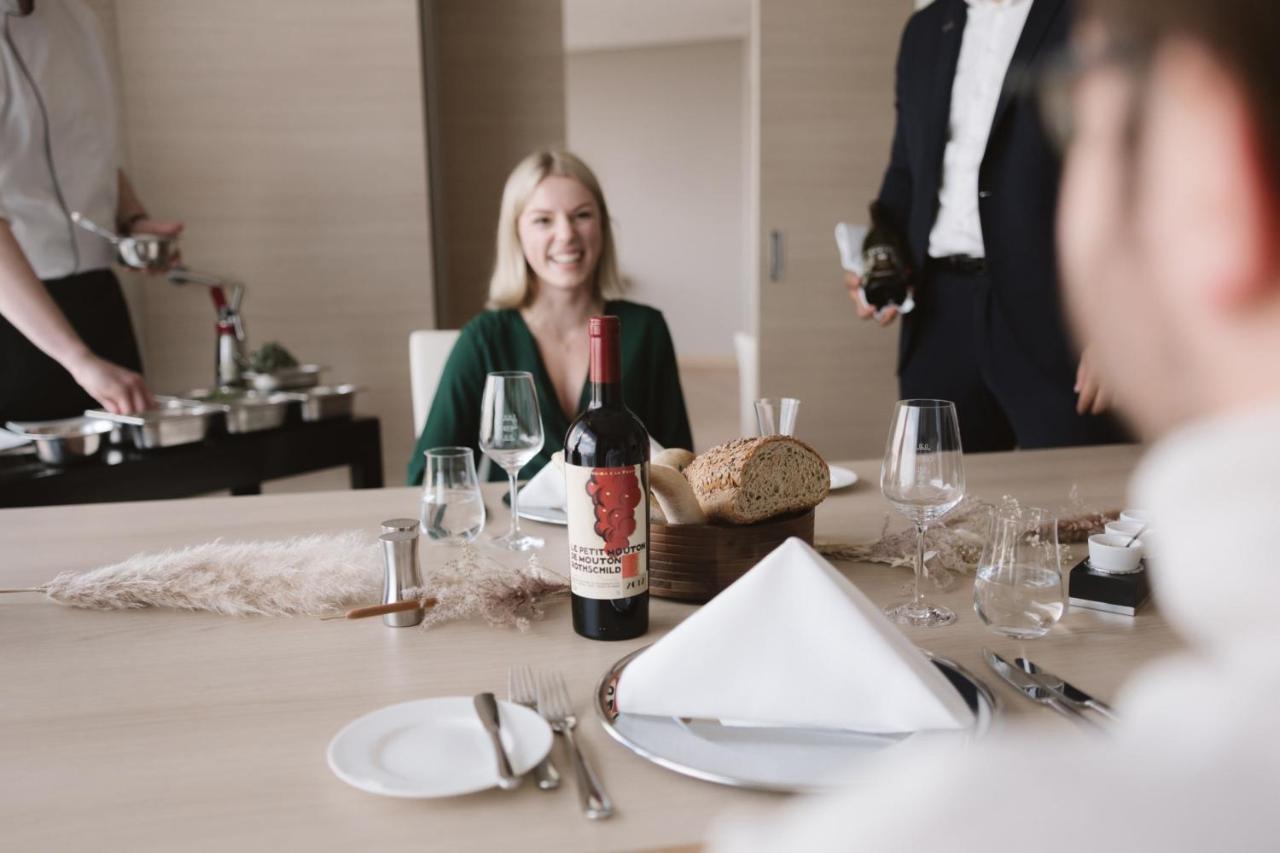 더 웨스틴 함부르크 함브루크 외부 사진 The image shows a dining setting with a woman smiling at the table. She is seated in front of a bottle of red wine that has a distinctive label. There is also bread and a neatly arranged table setting, including plates and glasses. In the background,