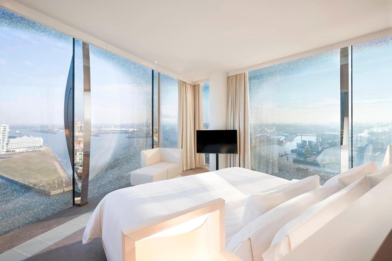 더 웨스틴 함부르크 함브루크 외부 사진 The photo shows a modern hotel room featuring a large bed with white linens and a minimalist design. It has expansive floor-to-ceiling windows that offer a panoramic view of the landscape outside, likely a city or waterfront setting. The room is deco