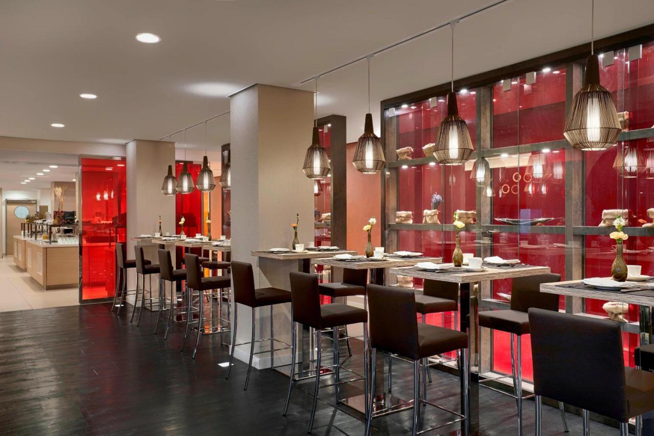 더 웨스틴 함부르크 함브루크 외부 사진 The photo shows a modern restaurant interior featuring a sleek, contemporary design. There are high tables with bar stools arranged along one side, illuminated by stylish hanging pendant lights. The color scheme includes warm tones, with red accents 