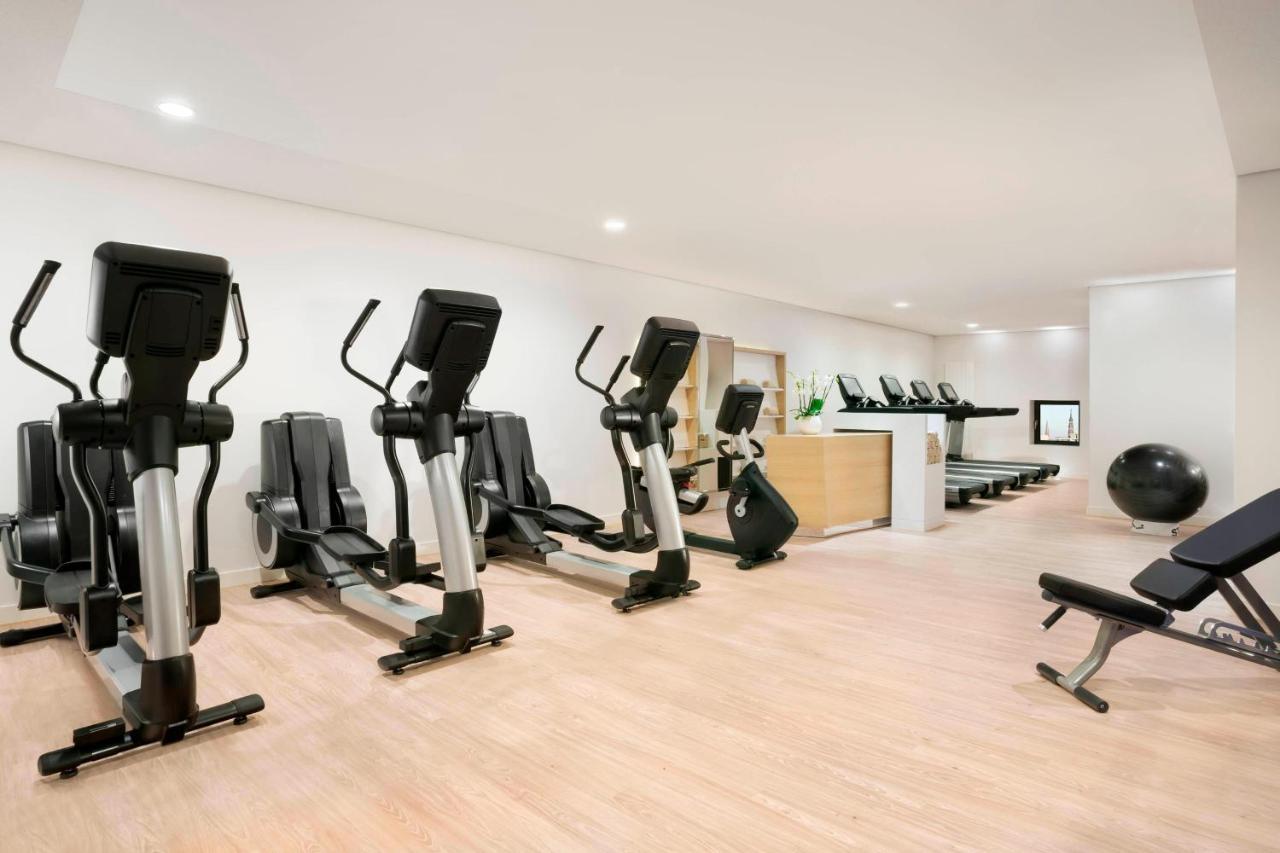 더 웨스틴 함부르크 함브루크 외부 사진 The photo shows a fitness room with several pieces of exercise equipment. On the left side, there are multiple elliptical machines arranged in a row. In the center, there is a stationary bike. A workout bench is positioned nearby, and a large exercis