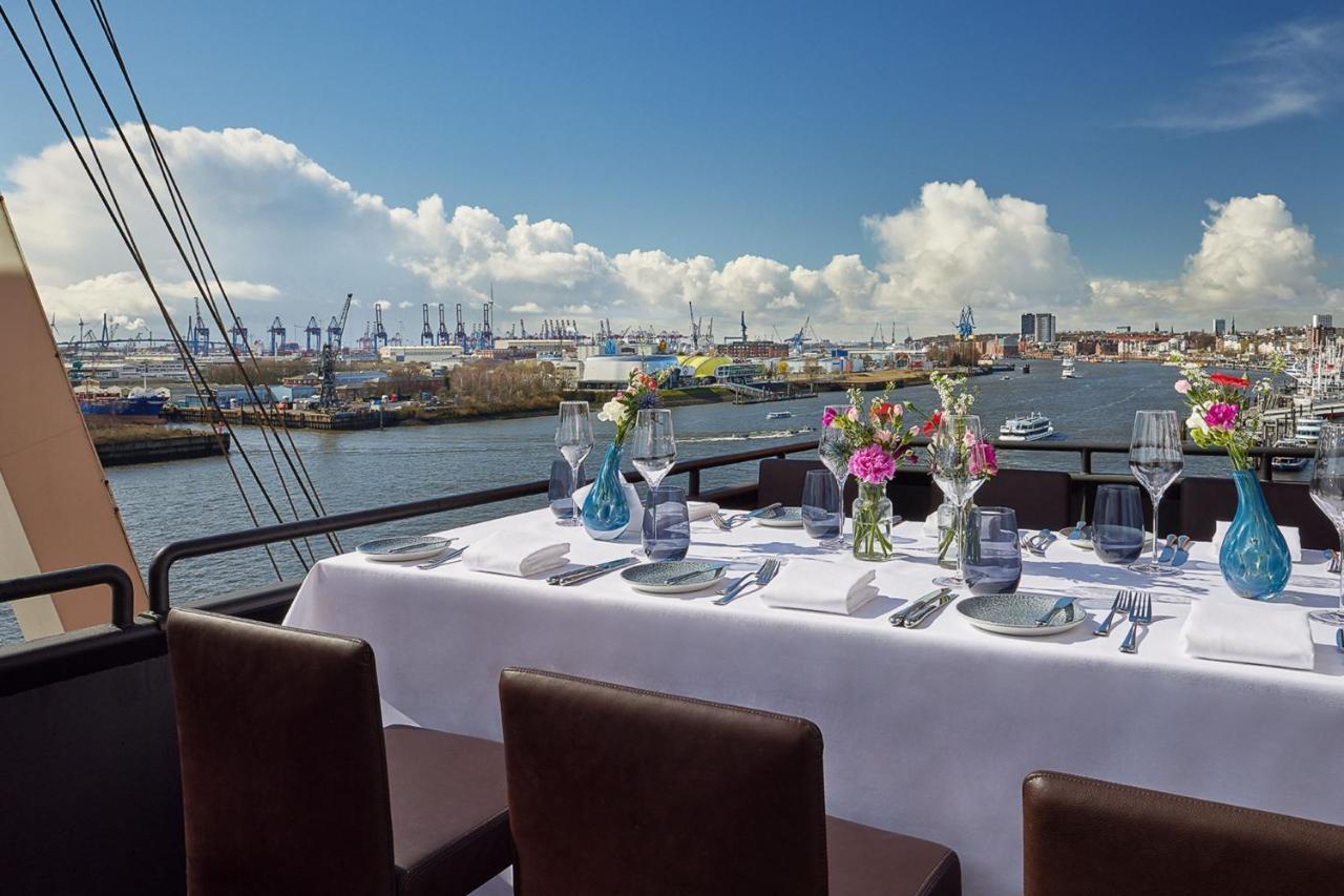 더 웨스틴 함부르크 함브루크 외부 사진 The photo shows a beautifully set dining table on a rooftop or terrace with a view of a body of water. The table is adorned with a white tablecloth, elegant tableware, and multiple vases filled with colorful flowers. In the background, there are boat
