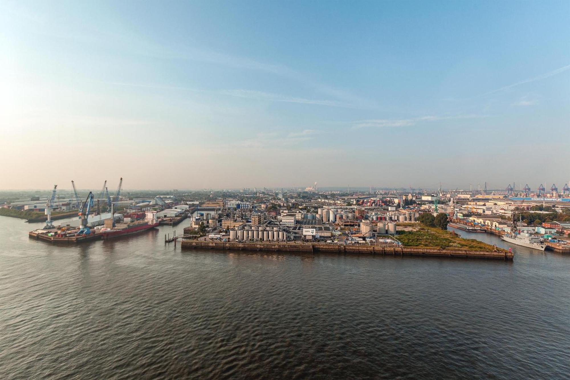 더 웨스틴 함부르크 함브루크 외부 사진 The photo shows a coastal area with a large body of water in the foreground. To the left, there are industrial structures like cranes and cargo ships, indicating an active port or shipping area. In the background, there are various buildings, which a