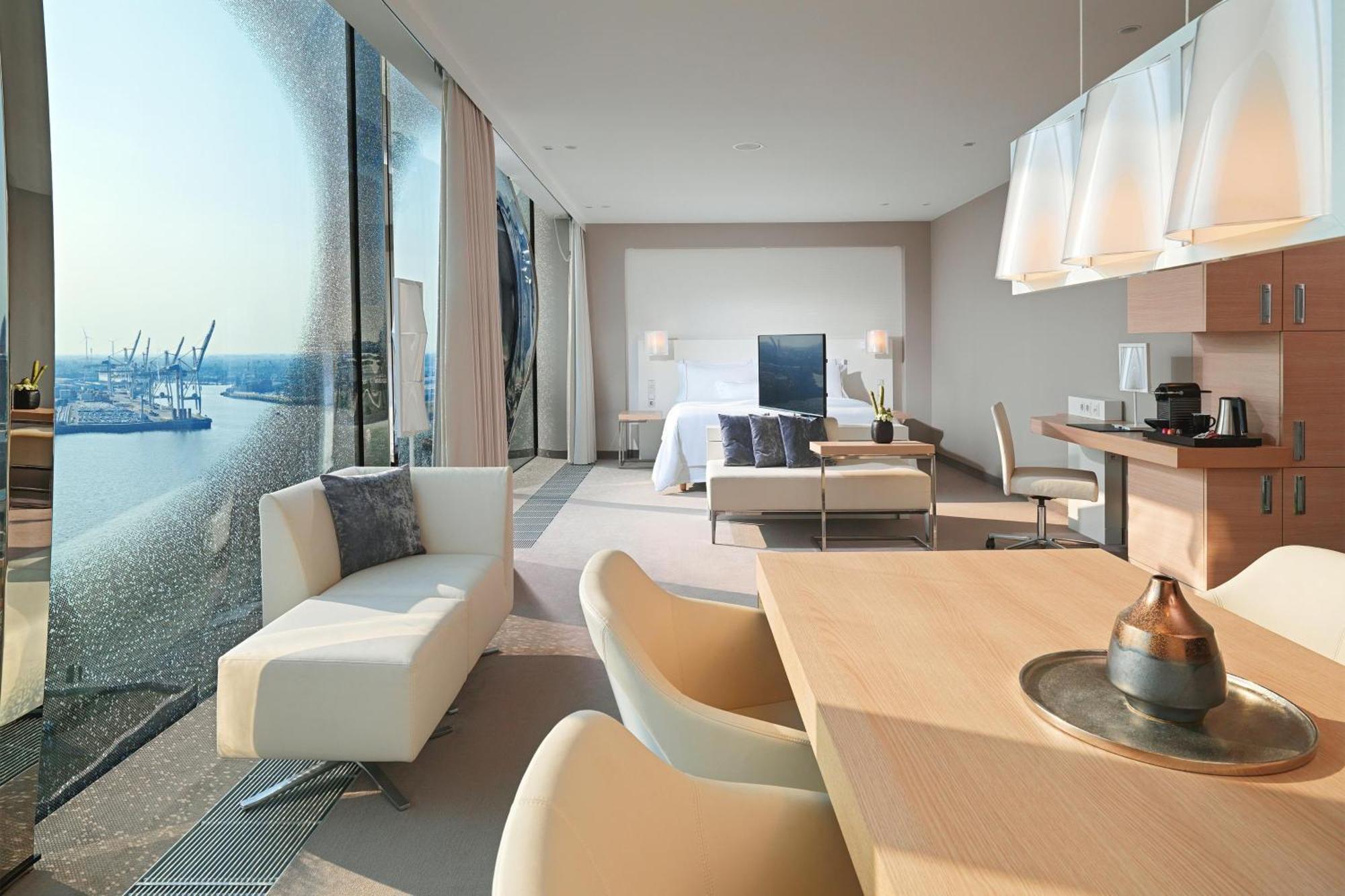 더 웨스틴 함부르크 함브루크 외부 사진 The photo shows a modern hotel room with a spacious layout. On one side, there are large floor-to-ceiling windows that offer a view of the outside, likely showcasing a waterfront or cityscape. Inside, there's a bed with a minimalist design, accompani
