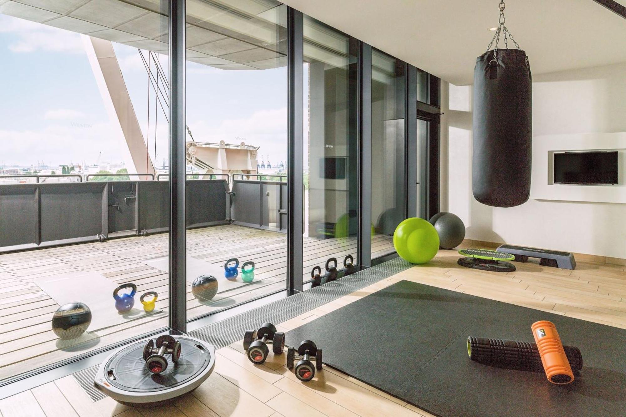 더 웨스틴 함부르크 함브루크 외부 사진 The photo shows a modern home gym with large glass windows that provide a view to the outside. Inside, there are various fitness equipment items, including a heavy punching bag hanging from the ceiling, kettlebells, a stability ball, an exercise mat,